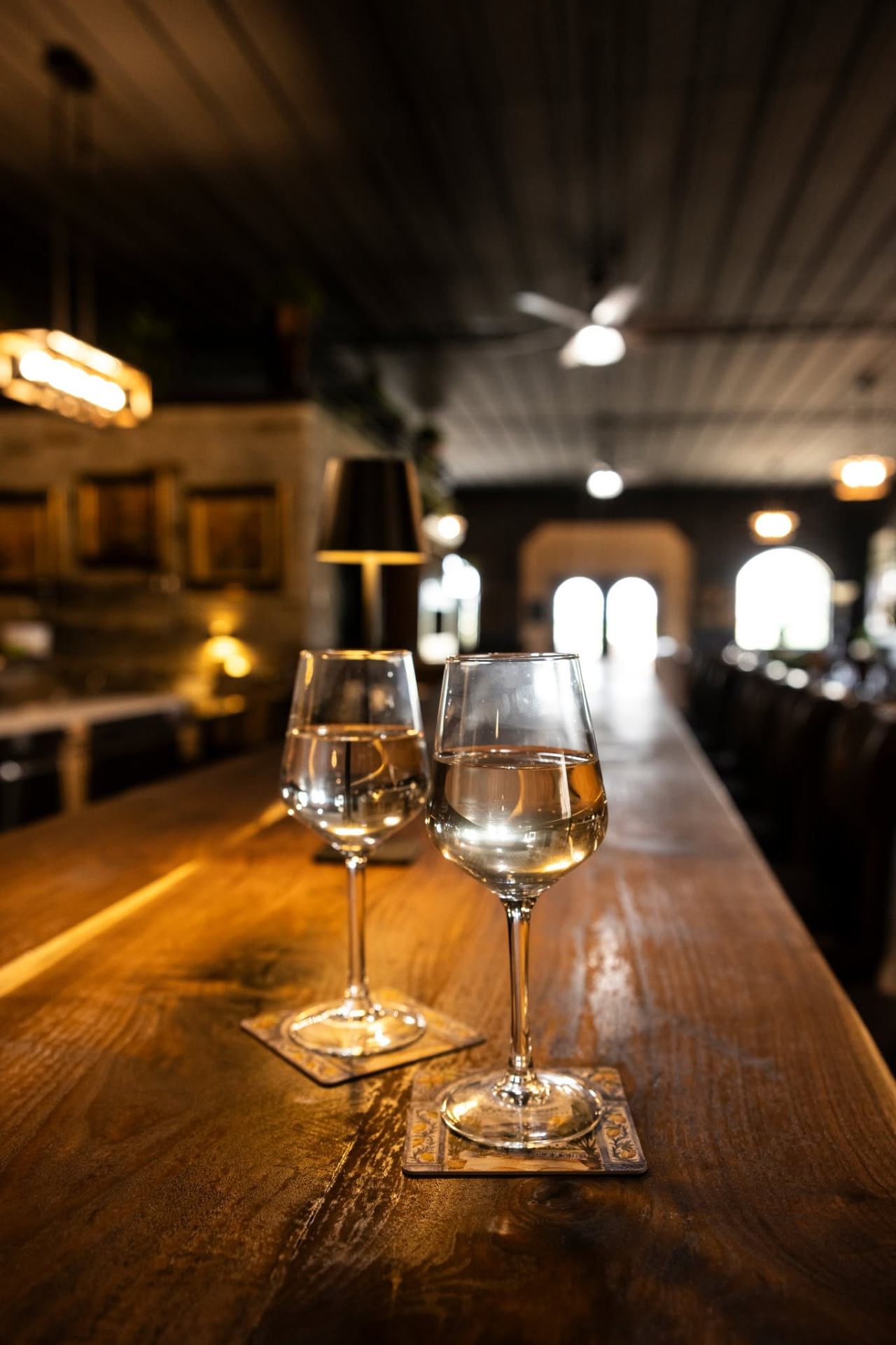 Two glasses of white wine on the bar with the hall glowing behind